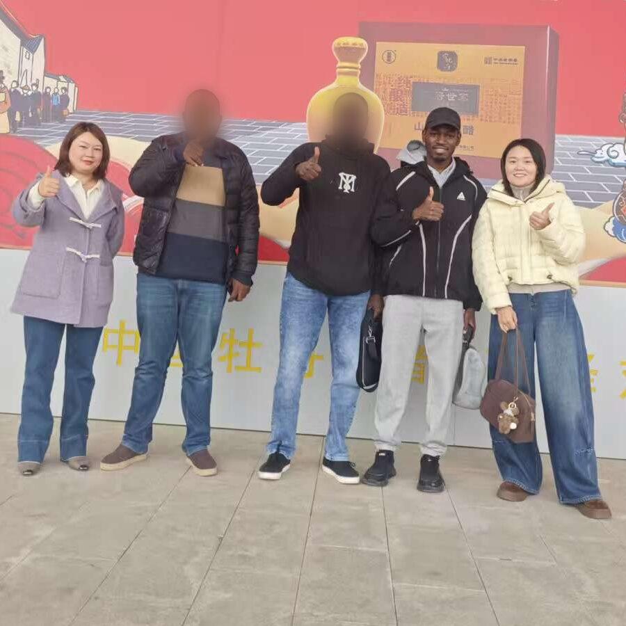 Youman with Nigerian customers and Chinese supplier staff in front of a factory banner. The customers' faces are blurred.